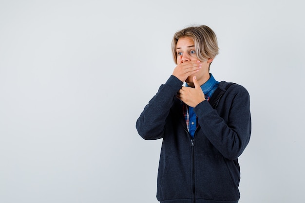 Ragazzo adolescente carino che tiene la mano sulla bocca in camicia, felpa con cappuccio e sembra spaventato. vista frontale.