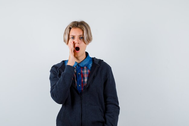 Ragazzo adolescente carino in camicia, felpa con cappuccio che tiene la mano vicino alla bocca e sembra agitato, vista frontale.