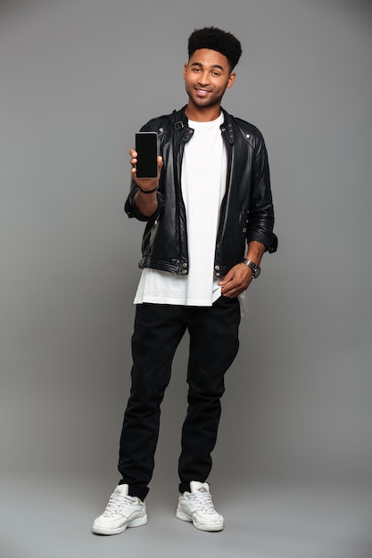 ragazzo africano alla moda sorridente in piedi con la mano in tasca mentre mostra schermo mobile vuoto, guardando