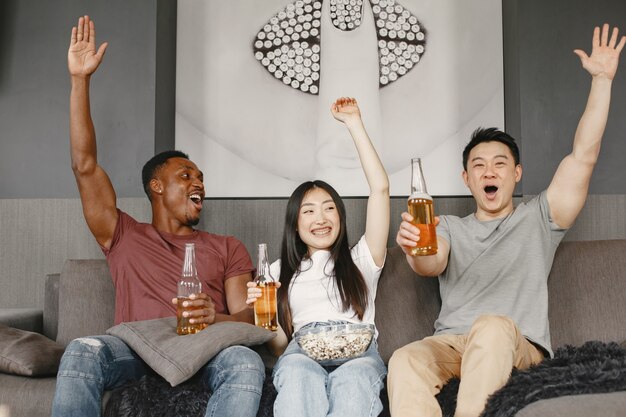 Ragazzo africano e coppia asiatica che guardano il calcio, mangiano popcorn e bevono birra. Amici che fanno il tifo per una squadra di calcio. Le persone sono felici.