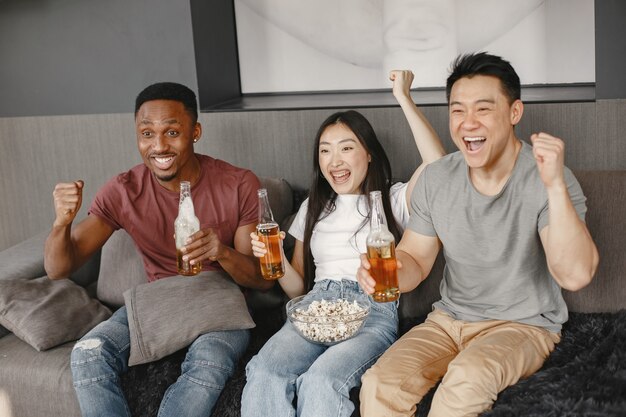 Ragazzo africano e coppia asiatica tintinnano una bottiglia con una birra. Amici che guardano la partita di calcio, mangiano popcorn. Persone che fanno il tifo per una squadra di calcio.