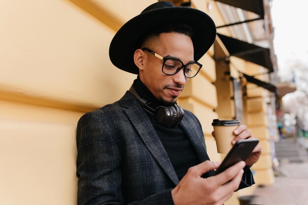 Ragazzo africano serio leggendo notizie in internet mentre beve il caffè. Foto all'aperto del giovane nero concentrato in cappello alla moda in piedi con il telefono e il latte vicino all'edificio.