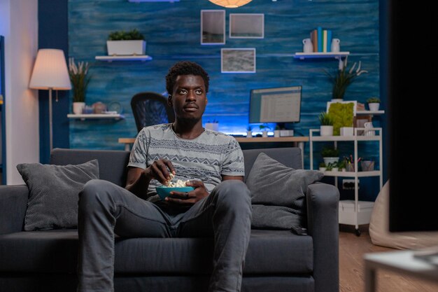 Ragazzo afroamericano che riposa sul divano in soggiorno tenendo una ciotola di popcorn mentre guarda un film di intrattenimento in televisione. Giovane che si diverte a passare il tempo libero da solo in soggiorno
