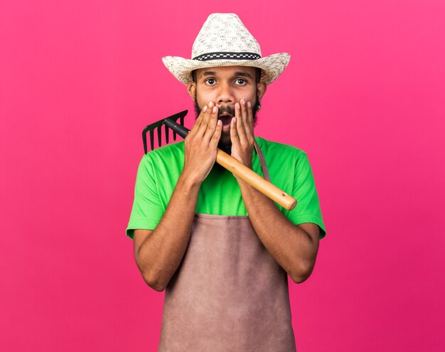 Ragazzo afroamericano del giovane giardiniere spaventato che indossa un cappello da giardinaggio che tiene la bocca coperta di rastrello con le mani isolate sul muro rosa