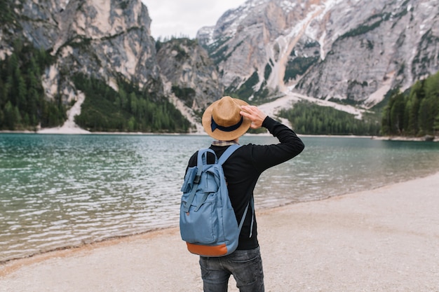 Ragazzo alla moda che indossa un cappello vintage decorato con nastro rilassante sulla riva del lago e guardando l'acqua