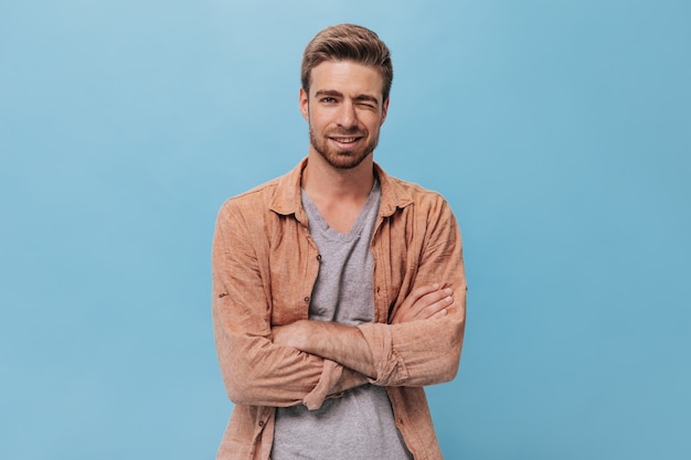 Ragazzo alla moda con la barba in camicia marrone e t-shirt sorridente e ammiccante sul muro isolato. Uomo figo in posa sul muro blu