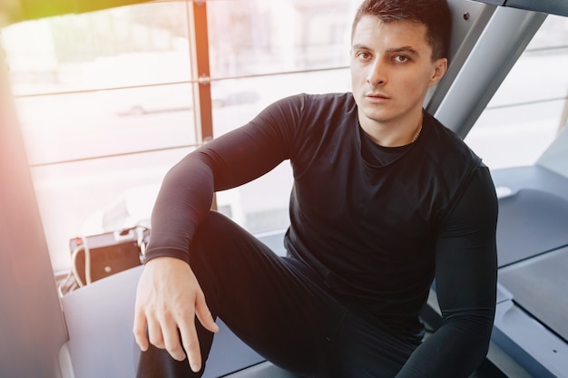 Ragazzo alla moda in palestra si siede a riposo sul tapis roulant. Uno stile di vita sano.