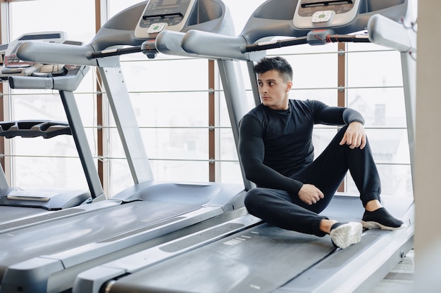 Ragazzo alla moda in palestra si siede a riposo sul tapis roulant. Uno stile di vita sano.