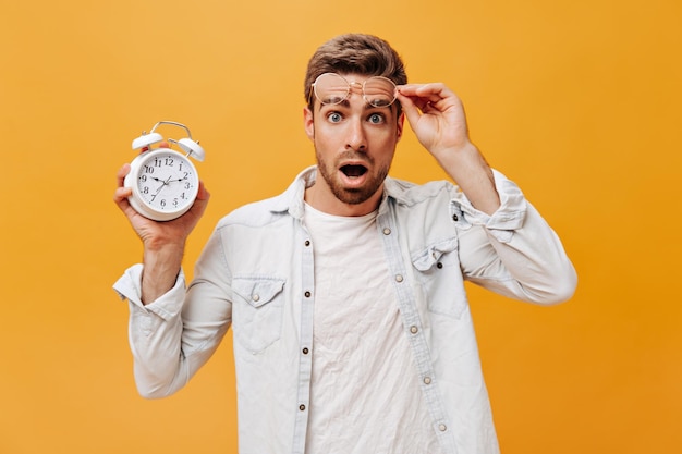 Ragazzo alla moda scioccato con gli occhi azzurri e la barba rossa in abito bianco che si toglie gli occhiali guardando nella fotocamera e tenendo l'orologio