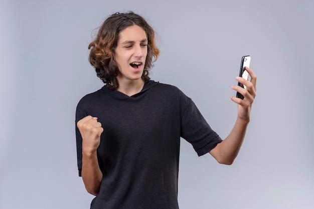 Ragazzo allegro con capelli lunghi in maglietta nera che tiene un telefono sul muro bianco