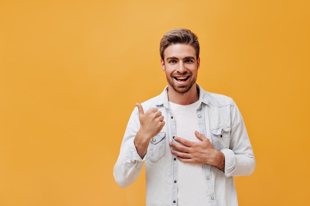 Ragazzo allegro con la barba in maglietta bianca e giacca di jeans che esamina la macchina fotografica e sorride su sfondo arancione isolato