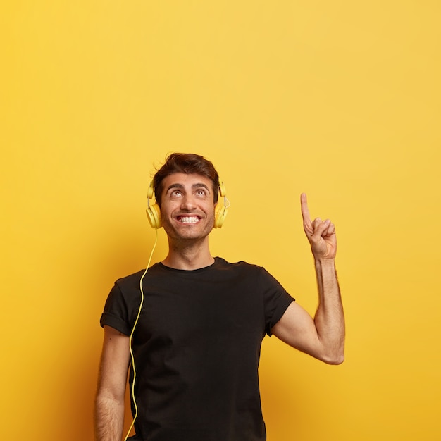 Ragazzo allegro con un sorriso a trentadue denti, punta il dito in alto, ascolta la musica in cuffia, gode di un buon suono