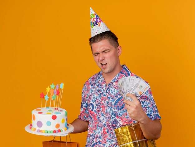 Ragazzo allegro del partito giovane con gli occhi chiusi che indossa il cappello di compleanno che tiene i regali con torta e contanti isolati sull'arancio