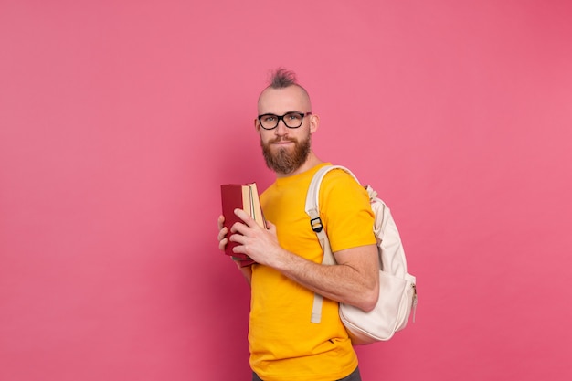 Ragazzo allegro di abbigliamento casual dell'allievo adulto con la barba e lo zaino che tengono i libri isolati sul colore rosa