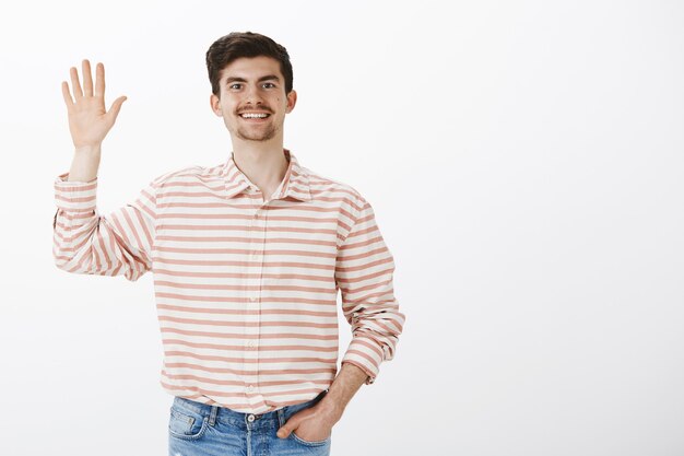 Ragazzo allegro e contento che dice ciao. Colpo di bel giovane ragazzo barbuto europeo con i baffi in camicia a righe, alzando la mano e salutando un amico, facendo un gesto di saluto, in piedi sopra il muro grigio