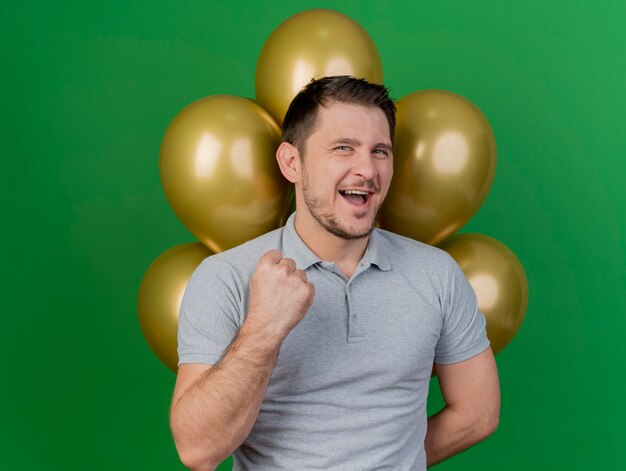 Ragazzo allegro festa giovane che indossa il cappello di compleanno in piedi davanti a palloncini e mostrando sì festure isolato sul verde