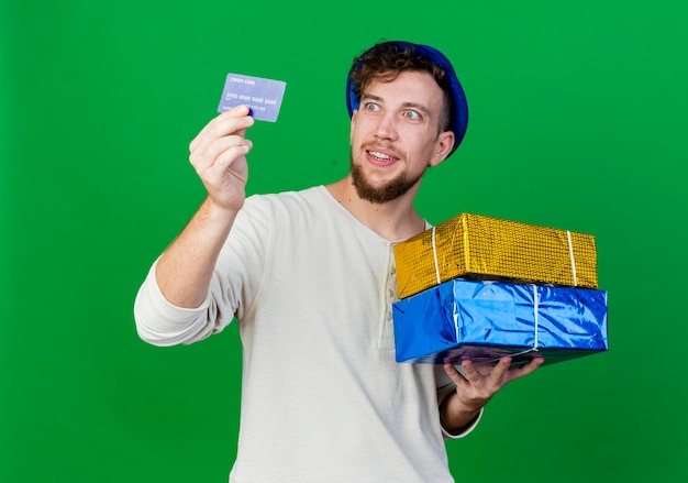 Ragazzo allegro giovane bello partito slavo che indossa il cappello del partito che tiene i contenitori di regalo e la carta di credito che esaminano la carta isolata sulla parete verde