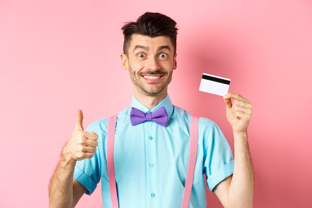 Ragazzo allegro in papillon che mostra pollice in su e carta di credito in plastica, come l'offerta promozionale, sorridendo felice alla telecamera, in piedi su sfondo rosa.