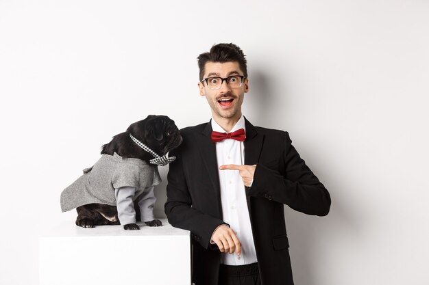 Ragazzo allegro in piedi con il suo carlino carino, puntando il dito contro il cane che indossa un costume da festa, in posa su sfondo bianco.