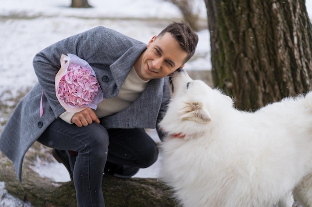 Ragazzo amico con un mazzo di fiori rosa ortensia che aspetta la sua ragazza e cammina e gioca con un cane. all'aperto mentre cade la neve. Concetto di San Valentino, proposta di matrimonio. l'uomo va