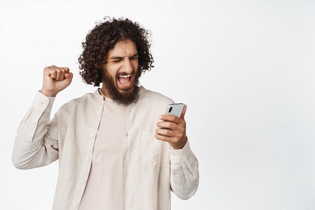 Ragazzo arabo allegro che guarda il telefono cellulare e festeggia guardando video di giochi sportivi su smartphone in piedi su sfondo bianco