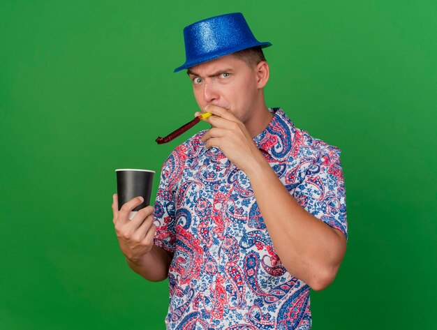 Ragazzo arrabbiato del partito giovane che indossa il cappello blu che tiene tazza di caffè e soffiante del partito di salto isolato su priorità bassa verde