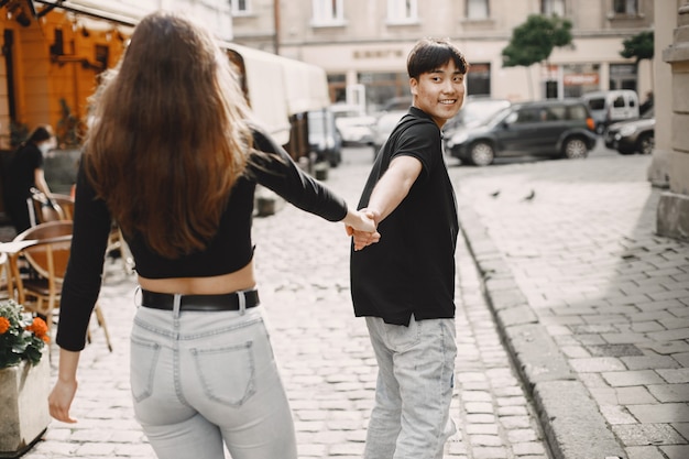 Ragazzo asiatico e la sua ragazza caucasica in abbigliamento casual camminando su un marciapiede sulla strada di Leopoli. Coppia che si tiene per mano mentre cammina insieme in città