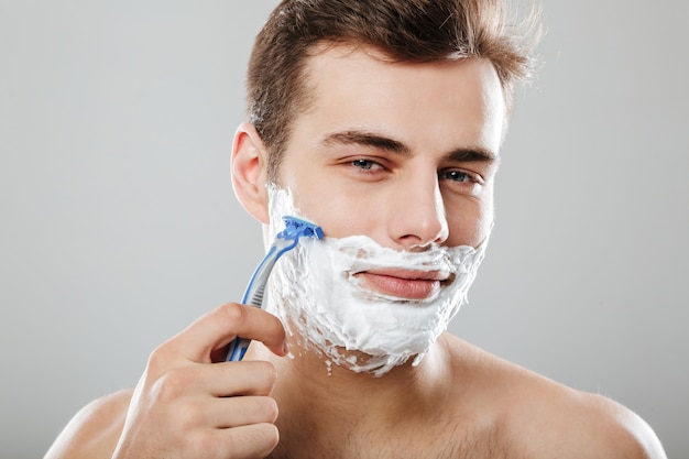 Ragazzo attraente con i capelli corti scuri che rade il suo fronte con il rasoio e il gel o la crema che sono isolati sopra la fine grigia della parete in su