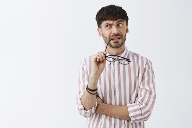 Ragazzo barbuto alla moda confuso e perplesso in posa contro il muro bianco con gli occhiali