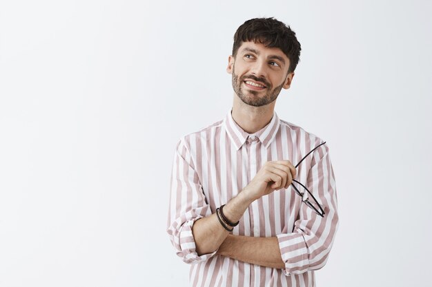 Ragazzo barbuto alla moda sognante e nostalgico che posa contro il muro bianco con gli occhiali