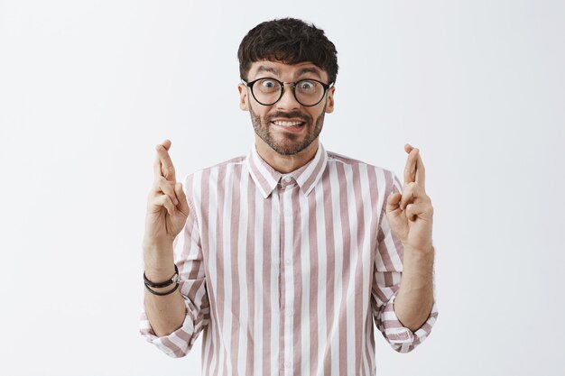 Ragazzo barbuto alla moda speranzoso che posa contro il muro bianco con gli occhiali