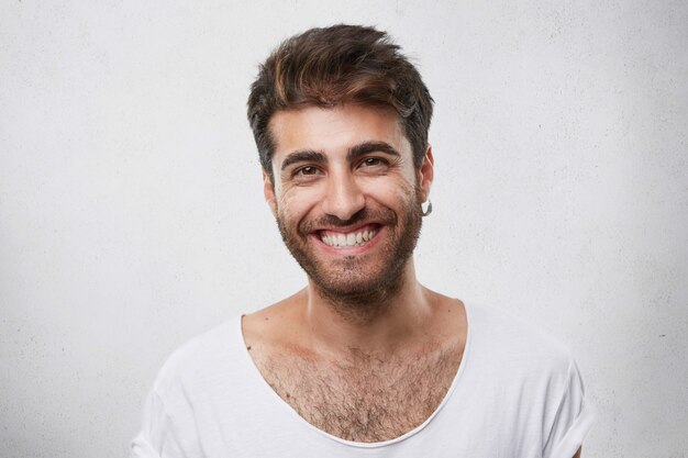 Ragazzo barbuto positivo con occhi scuri e caldi con orecchino nell'orecchio vestito casualmente con un sorriso piacevole che mostra i suoi denti bianchi perfetti che hanno buon umore