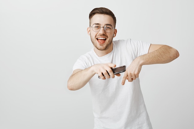 Ragazzo bello felice con gli occhiali ballare la musica in auricolari wireless con il cellulare in mano