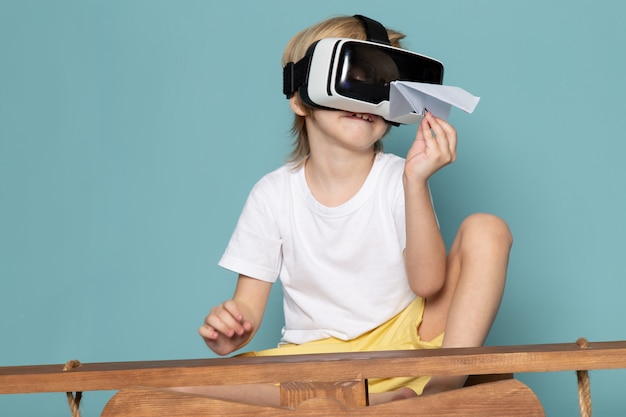 ragazzo biondo di vista frontale che gioca gli occhiali di protezione del vr che tengono aereo di carta sull'azzurro