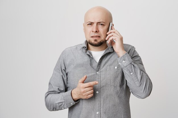 Ragazzo calvo di mezza età incerto che strizza gli occhi perplesso, conversando al telefono