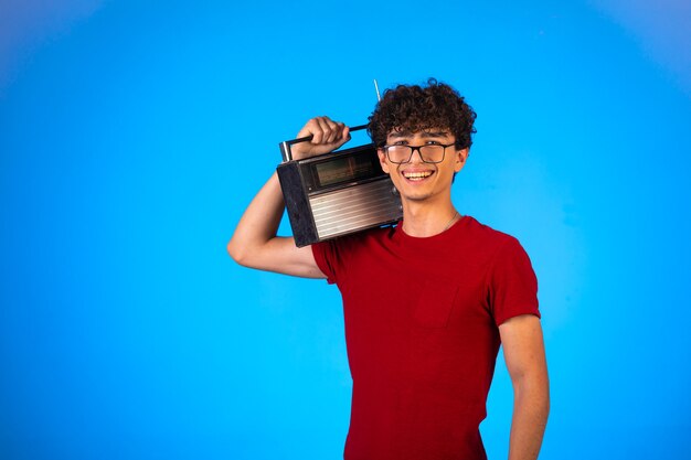 Ragazzo capelli ricci in camicia rossa che tiene una radio d'epoca e sorridente.