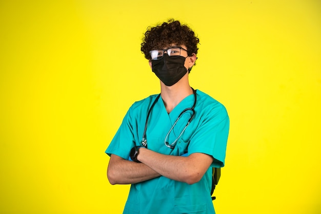 Ragazzo capelli ricci in uniforme medica e maschera facciale in piedi in una posizione sicura.