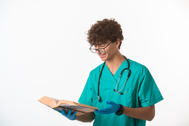 Ragazzo capelli ricci in uniforme medica e maschere per le mani leggendo un vecchio libro e sorridente.