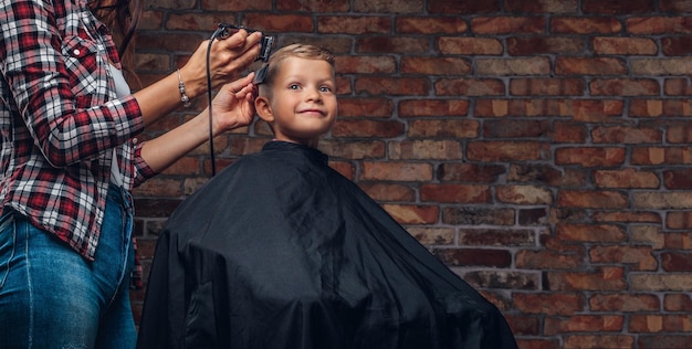 Ragazzo carino bambino in età prescolare che ottiene taglio di capelli. Il parrucchiere per bambini con il rifinitore sta tagliando il ragazzino nella stanza con interni soppalcati.