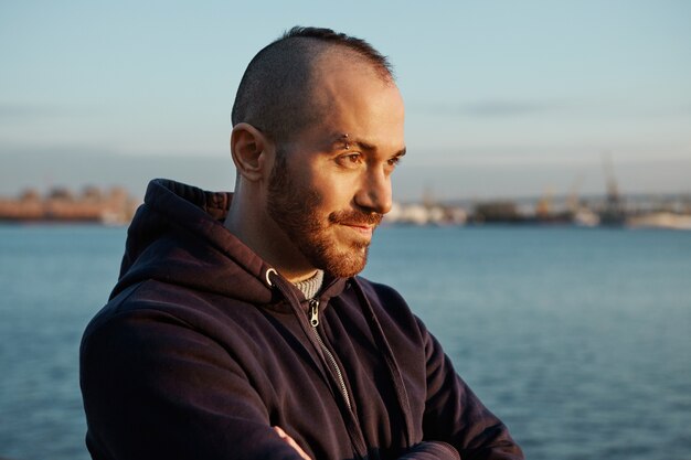 Ragazzo carino hipster con sopracciglio trafitto, barba tagliata e acconciatura alla moda ammirando la vista sul fiume