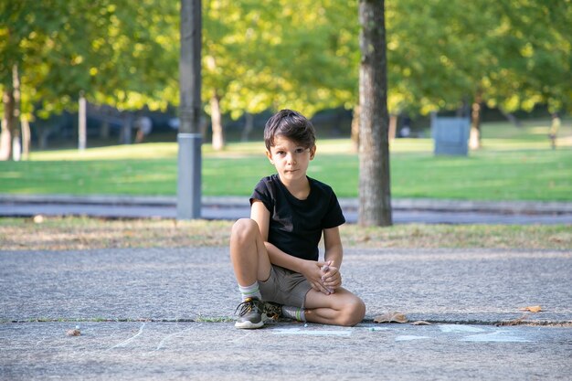 Ragazzo carino serio seduto e disegno su asfalto con pezzi colorati di gessi. Vista frontale. Concetto di infanzia e creatività