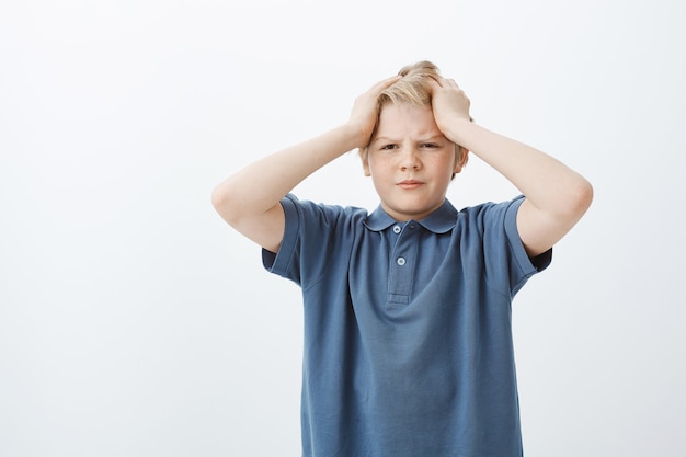 ragazzo carino triste e turbato con i capelli biondi, tenendo le mani sulla testa e guardando con espressione dispiaciuta da parte