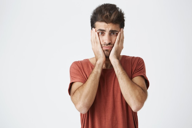 Ragazzo caucasico attraente maturo in maglietta rossa e acconciatura alla moda tenendo il viso con le mani sconvolte per una grande quantità di lavoro.