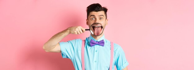Ragazzo caucasico divertente che mostra la lingua nella lente d'ingrandimento sorridendo e facendo facce in piedi in cravatta a farfalla o