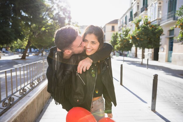 Ragazzo che bacia giovane signora sorridente con palloncini sulla strada