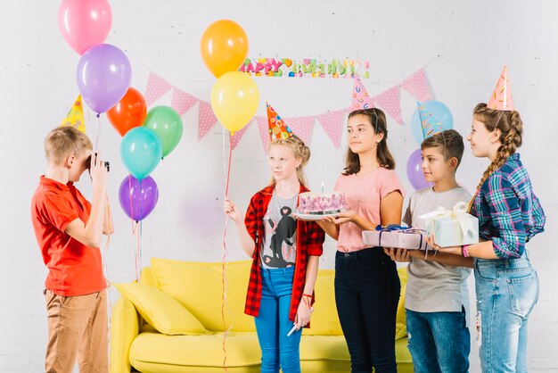 Ragazzo che cattura foto dei suoi amici con torta di compleanno; regalo e palloncini