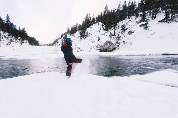 Ragazzo che dà dei calci alla neve sulla riva del fiume