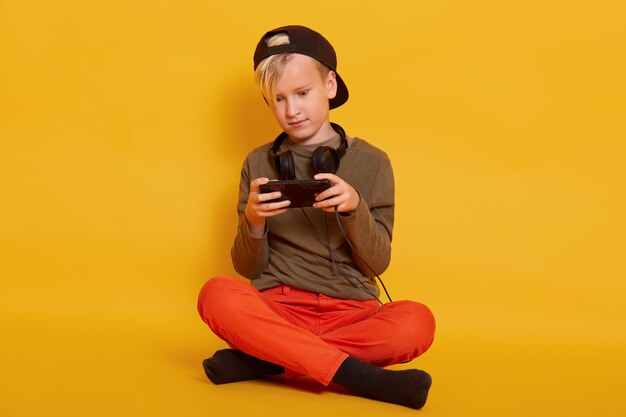 Ragazzo che gioca tramite telefono cellulare, adorabile bambino maschio seduto isolato su giallo e tenendo mobile, ragazzo si veste casualmente, in posa con le cuffie intorno al collo, mantenendo le gambe incrociate.