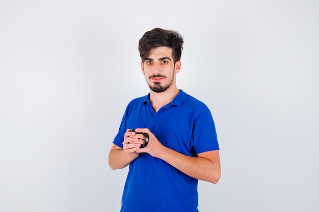 Ragazzo che tiene tazza con entrambe le mani in maglietta blu e sembra serio. vista frontale.