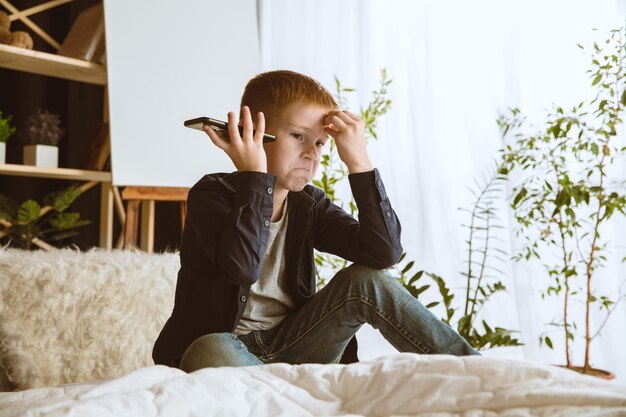 Ragazzo che utilizza diversi gadget a casa. Piccolo modello con smartwatch, smartphone o tablet e auricolari. Fare selfie, chattare, giocare, guardare video. Interazione di bambini e moderne tecnologie.